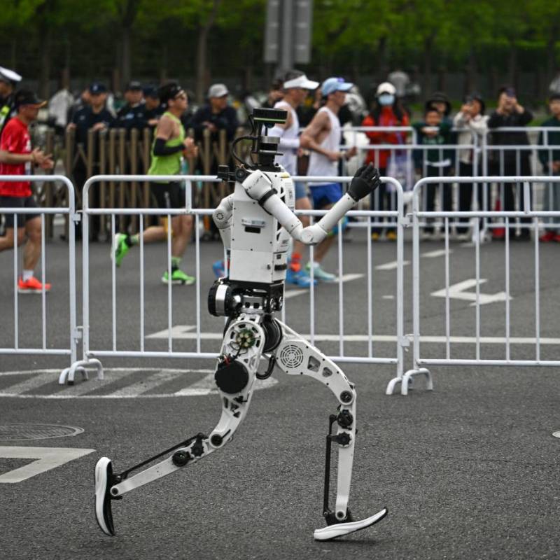 En un carril, uno de los robots humanoides participantes, en el otro, las personas corriendo la media maratón de Pekín. FOTO AFP. 