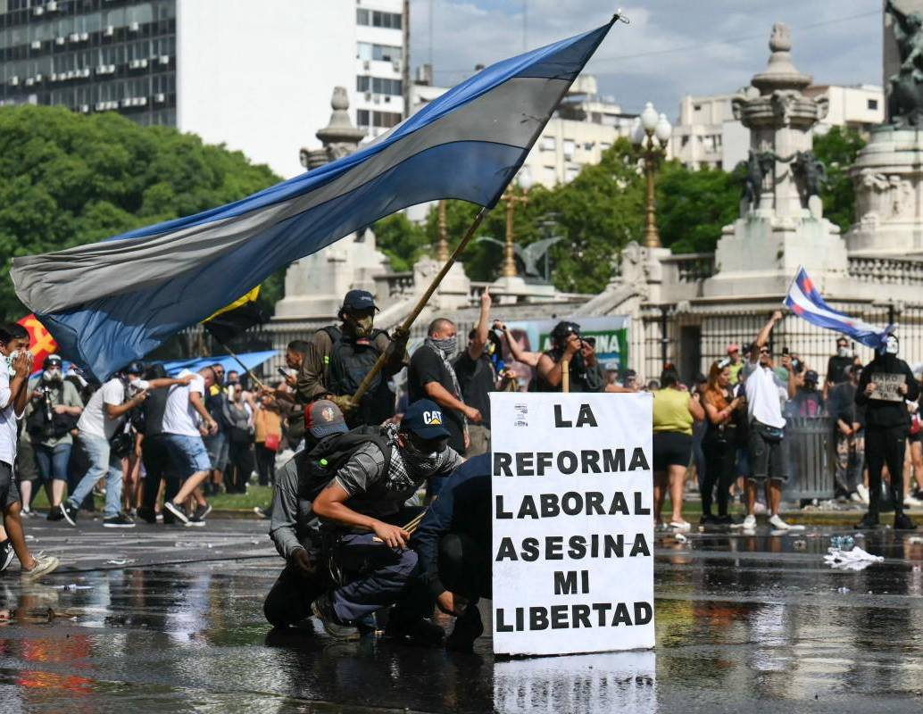 Manifestantes se protegen tras una pancarta que dice “La reforma laboral mata mi libertad” de un cañón de agua disparado por la policía antidisturbios durante una protesta convocada por sindicalistas contra el debate sobre la reforma laboral en el Congreso Nacional en Buenos Aires. FOTO: AFP.