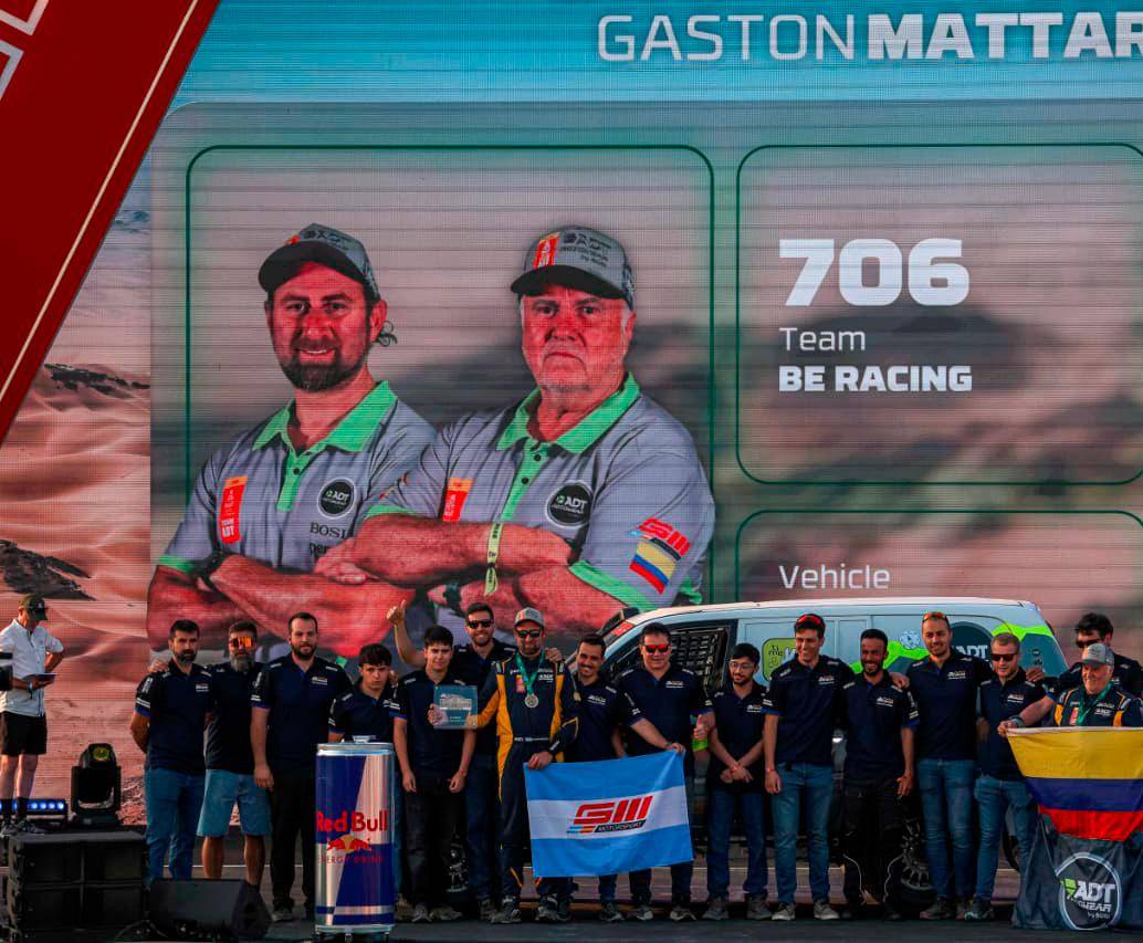 En la imagen aparece el colombiano Javier Vélez, en la pantalla, con gorra gris y barba canosa, y abajo sosteniendo la bandera de Colombia durante el festejo de su equipo en el Rally Dakar. <span class="mln_uppercase_mln">FOTO</span> <b><span class="mln_uppercase_mln">cortesía adt motowear</span></b>