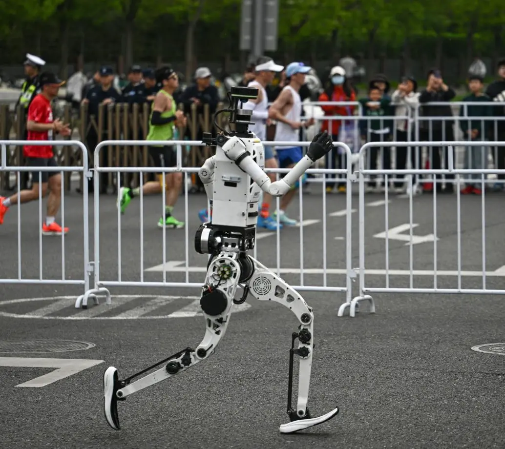 En un carril, uno de los robots humanoides participantes, en el otro, las personas corriendo la media maratón de Pekín. FOTO AFP. 