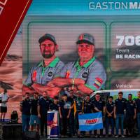 En la imagen aparece el colombiano Javier Vélez, en la pantalla, con gorra gris y barba canosa, y abajo sosteniendo la bandera de Colombia durante el festejo de su equipo en el Rally Dakar. FOTO cortesía adt motowear