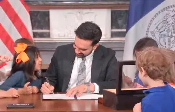 El alcalde de Nueva York, Zohran Mamdani, firma la orden ejecutiva simbólica que “deroga” la hora de dormir de los escolares para las Finales de la NBA, acompañado por niños que estamparon sus huellas en el documento. Foto: Captura de video