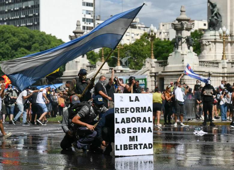 Manifestantes se protegen tras una pancarta que dice “La reforma laboral mata mi libertad” de un cañón de agua disparado por la policía antidisturbios durante una protesta convocada por sindicalistas contra el debate sobre la reforma laboral en el Congreso Nacional en Buenos Aires. FOTO: AFP.