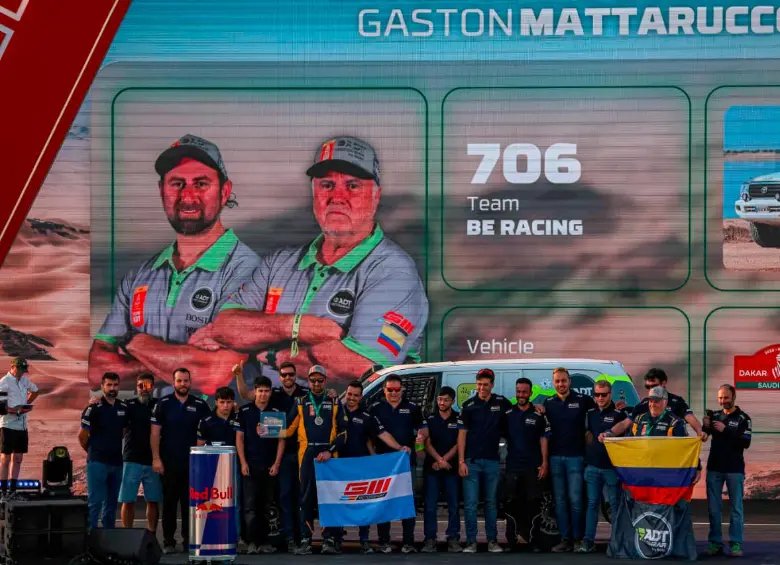 En la imagen aparece el colombiano Javier Vélez, en la pantalla, con gorra gris y barba canosa, y abajo sosteniendo la bandera de Colombia durante el festejo de su equipo en el Rally Dakar. FOTO cortesía adt motowear