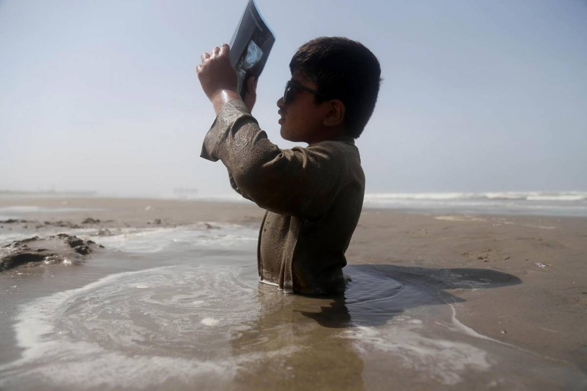 Un niño con discapacidad está sentado a medio enterrar en la orilla del río Indo, y utiliza una placa de rayos X para observar un eclipse solar parcial, siguiendo el consejo de un médico de permanecer en la arena durante el suceso. FOTO EFE