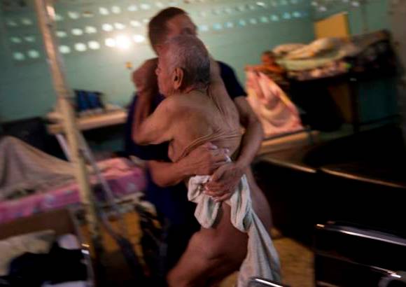 Reuters - Danny Martínez, un paciente en rehabilitación de drogas, ayuda a pasar a un anciano de su silla de ruedas a la cama, después de bañarlo en el centro de rehabilitación para adictos a las drogas y el alcohol Nosotros Unidos en Caracas. Este centro está financiado por una iglesia cristiana, y ha sido un modelo para las instituciones del Estado con la mismo objetivo.