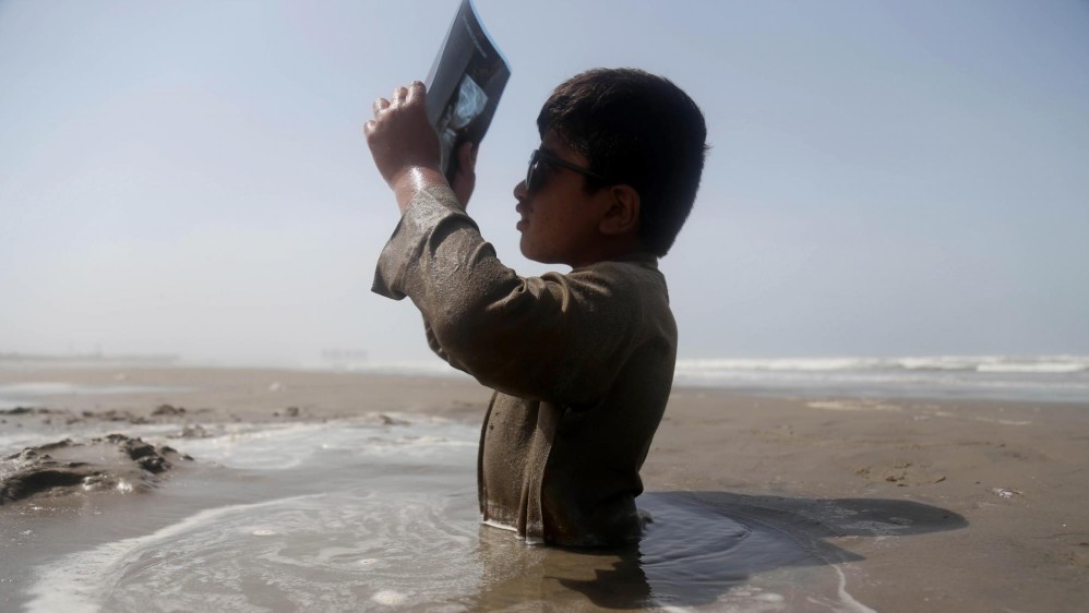 Un niño con discapacidad está sentado a medio enterrar en la orilla del río Indo, y utiliza una placa de rayos X para observar un eclipse solar parcial, siguiendo el consejo de un médico de permanecer en la arena durante el suceso. FOTO EFE